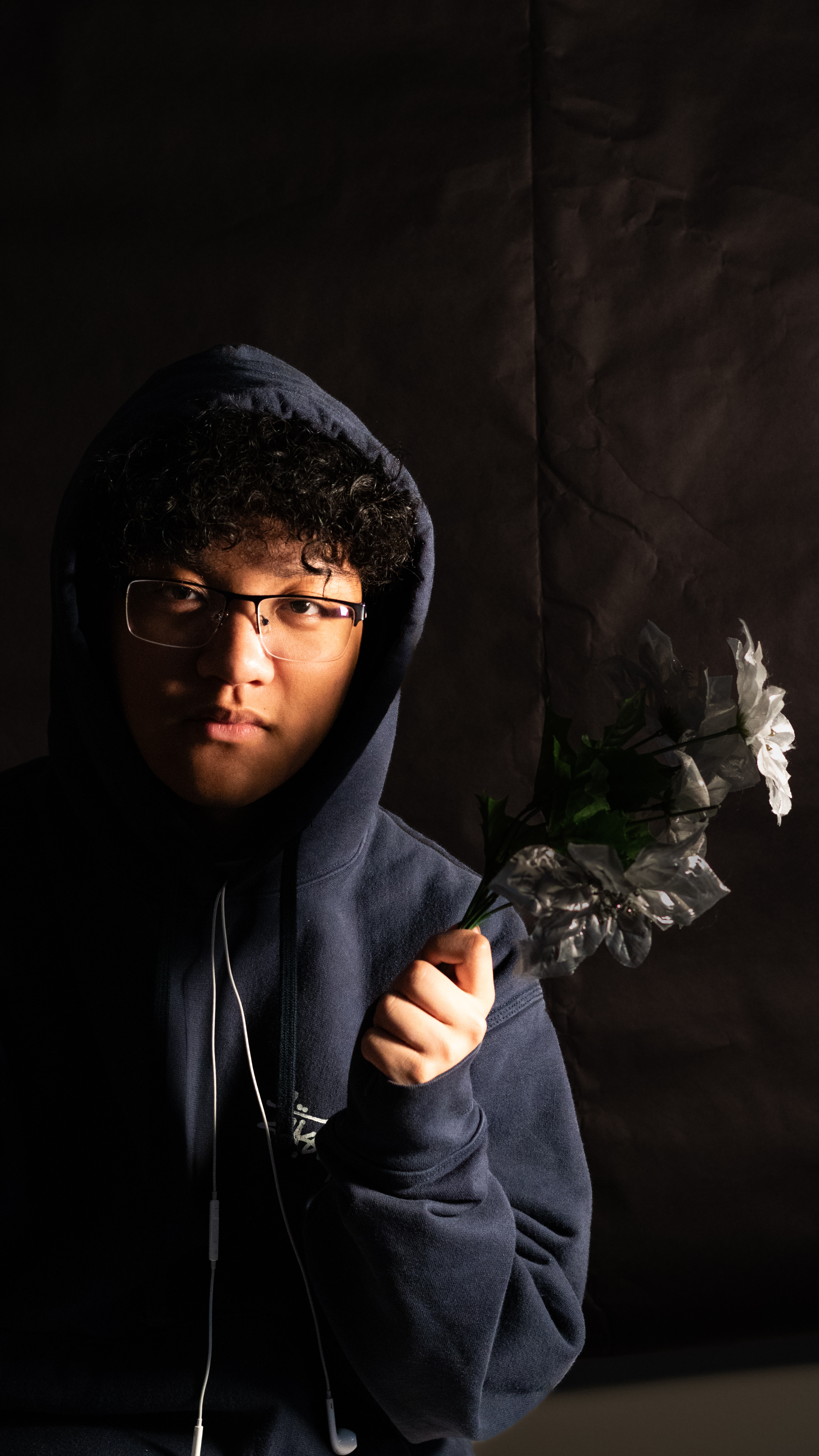 An expressionless male face against a black background. A glint of light shines off his face, and he his holding a bunch of white flowers in his hand.